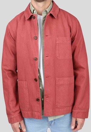 Homme portant une veste rouge déboutonnée sur une chemise verte et un t-shirt blanc, avec un jean bleu clair, debout devant un fond uni.