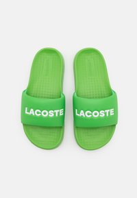 Sandales en caoutchouc vertes avec de larges brides portant le logo "LACOSTE" en blanc. Semelle intérieure texturée et surface supérieure lisse.