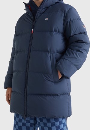 Osoba nosící tmavě modrou prošívanou bundu s kapucí, zipem, bočními kapsami a malými logy na hrudi a rukávu, v kombinaci s modrými károvanými kalhotami.