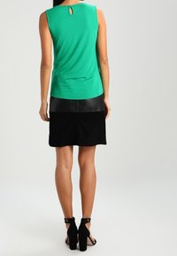 Top vert sans manches avec une ouverture dans le dos, associé à une jupe noire. La jupe présente un mélange de textures, lisses et douces, avec des chaussures à talons empilés.