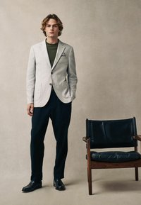 Jasnoszary lniany blazer z klapami, zielona koszula pod spodem, ciemne spodnie i czarne mokasyny. Drewniane krzesło z czarnym skórzanym siedziskiem.