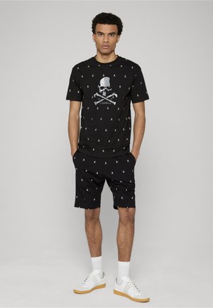 Mand stående med hænderne i lommerne iført sort t-shirt med kranieprint og matchende shorts med hvide sokker og hvide sneakers.