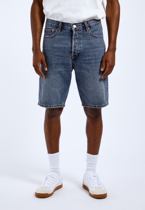 Mand iført midilengde blå denimshorts, hvide sokker, hvide sneakers og en hvid skjorte, stående foran en ensfarvet baggrund.