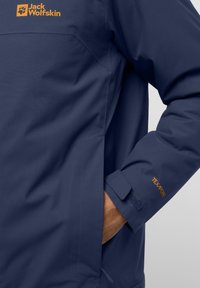 Veste imperméable marine avec un tissu texturé, poignet ajustable avec velcro et une étiquette de logo. Comprend une poche et un design épuré.