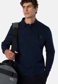 Camisa polo de manga larga en color marino con una tela texturizada, tapeta de botones y un bolsillo en el pecho. Sosteniendo una bolsa de deporte gris con acentos rojos.