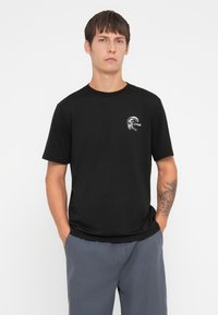 Sort bomulds t-shirt med et lille hvidt grafisk logo på brystet. Modellen har grå shorts på, hænderne i lommerne, med en enkel baggrund.