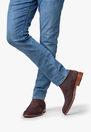 Botas de ante marrón hasta el tobillo con paneles laterales elásticos y tacón de madera, combinadas con unos jeans azules ajustados, mostrando un look casual pero refinado.
