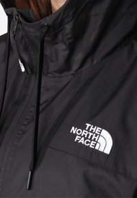 Giacca impermeabile nera con colletto alto, coulisse regolabili e logo ricamato bianco sul petto. Tessuto liscio.