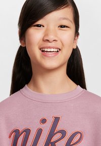 Sudadera morada con cuello acanalado que presenta un destacado logotipo bordado de "nike" en azul marino y naranja. La persona que la lleva tiene el pelo largo y negro.