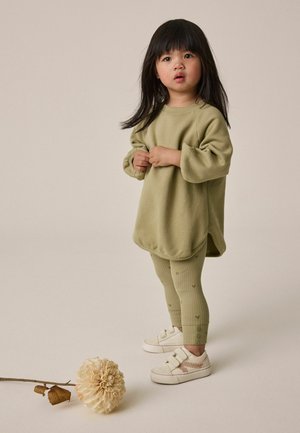 Jong kind met lang donker haar, gekleed in een te grote groene trui, groene legging met patroon en witte sneakers, staat dichtbij een grote crèmekleurige bloem op de vloer.
