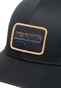 Svart caps laget av teksturert stoff med en rektangulær lapp som har "BILLABONG" i flerfarget broderi og en gyllen kant.