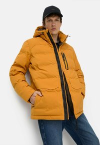 Orange pufferjakke med lynlås foran, store lommer og en sort accentlynlås; sat sammen med mørke jeans og en sort kasket.