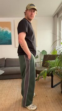 Zielone spodnie dresowe z białymi bocznymi paskami, czarna koszulka i beżowa czapka. Stojąca poza z rękami w kieszeniach, w pomieszczeniu. Lekka faktura i luźny krój.