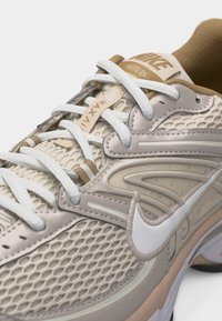 Μπεζ και άσπρο αθλητικό παπούτσι Nike για τρέξιμο με επάνω μέρος από διχτυωτό υλικό, επενδεδυμένο γιακά, λευκό λογότυπο swoosh και λευκά κορδόνια δεμένα σε ουδέτερο φόντο.