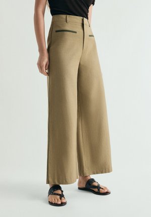 Vrouw draagt beige, hoog getailleerde, brede broek met zwarte nep-achterzakken en zwarte gespen sandalen met studs, staand tegen een effen achtergrond.