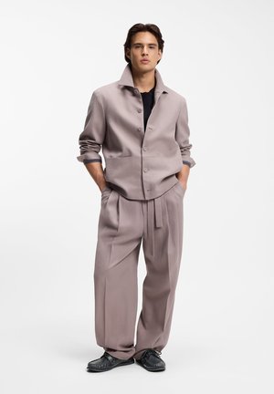 Giovane uomo in piedi con le mani nelle tasche, indossa una giacca larga color malva chiaro e pantaloni abbinati a gamba larga, con scarpe nere su sfondo bianco.