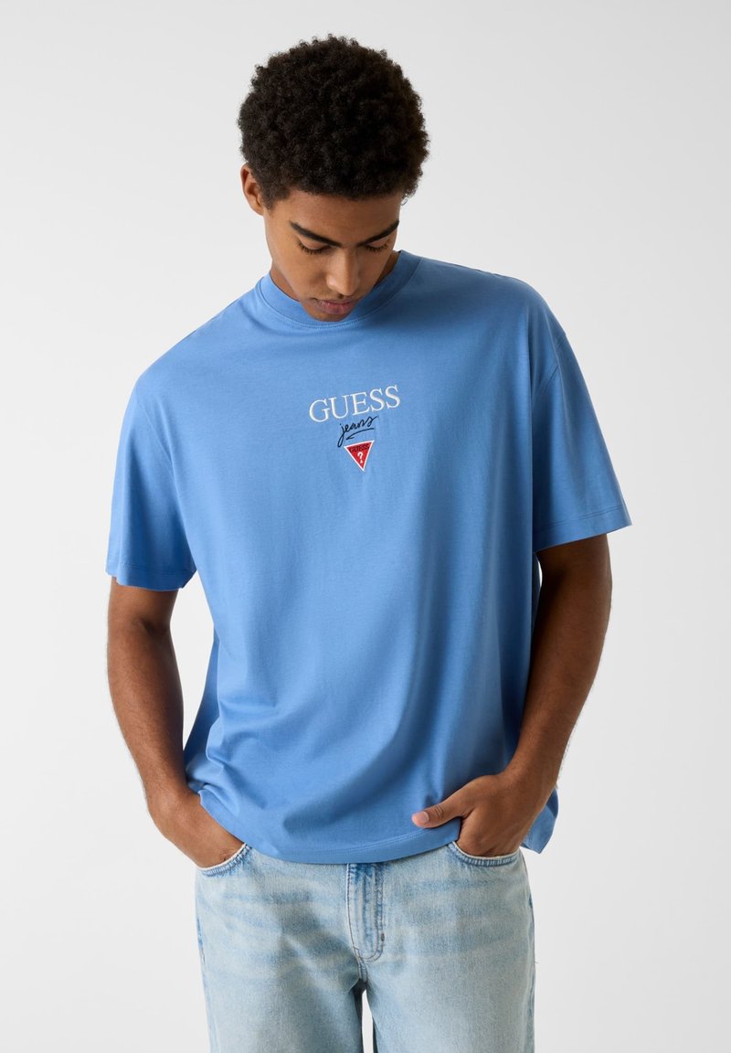 Φωτεινό γαλάζιο βαμβακερό t-shirt με κοντά μανίκια, με λευκό λογότυπο "GUESS jeans". Κλασικός στρογγυλός λαιμός και άνετη εφαρμογή.