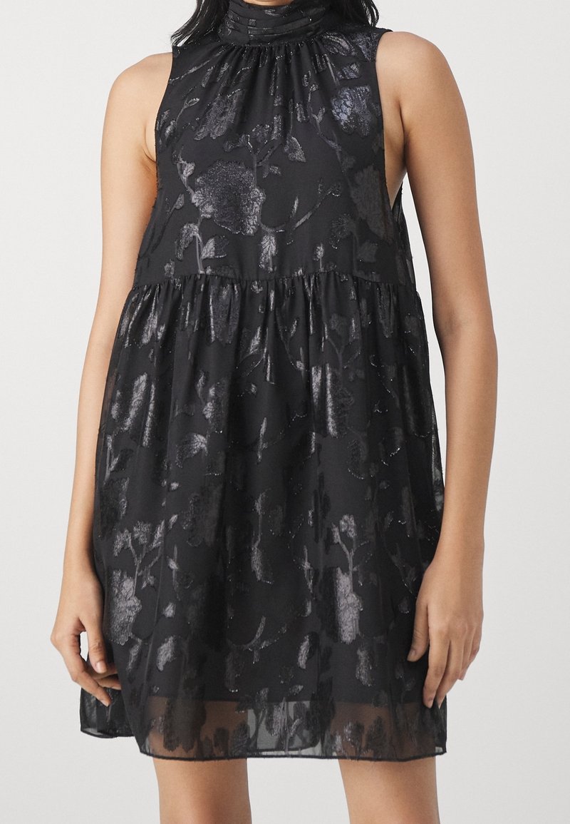 Robe noire sans manches avec col montant, coupe ample, et motif floral brillant, portée par une personne debout les bras détendus le long du corps.