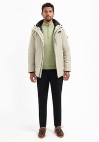 Beige geïsoleerde jas met een zwarte capuchon, groene pullover eronder, zwarte broek en bruine veterschoenen met een gestructureerde zool.