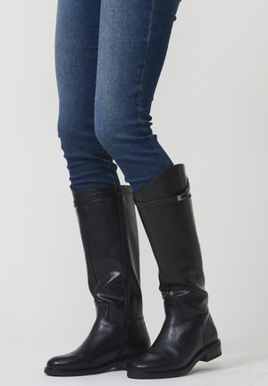 Persona vestida con jeans ajustados azul oscuro y botas de cuero negras hasta la rodilla con cremalleras laterales y tacones bajos, de pie sobre un fondo liso.