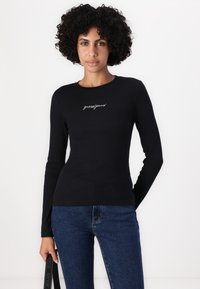 Juodas ilgų rankovių marškinėliai su raštuota tekstūra, apvaliu iškirpimu ir "guessjeans" siuvinėjimu ant krūtinės. Derinama su mėlynais džinsais.