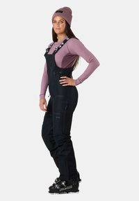 Pantaloni neri impermeabili a salopette con spalline regolabili, tasche con zip e design aderente, indossati con una maglietta a maniche lunghe viola chiaro e un berretto.