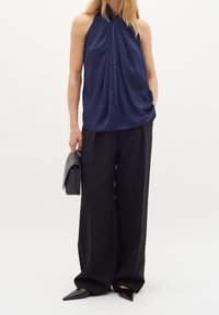 Mörkblå ärmlös topp med samlad detalj vid halsen, kombinerad med svarta vida byxor och spetsiga svarta skor, hållande en svart handväska.