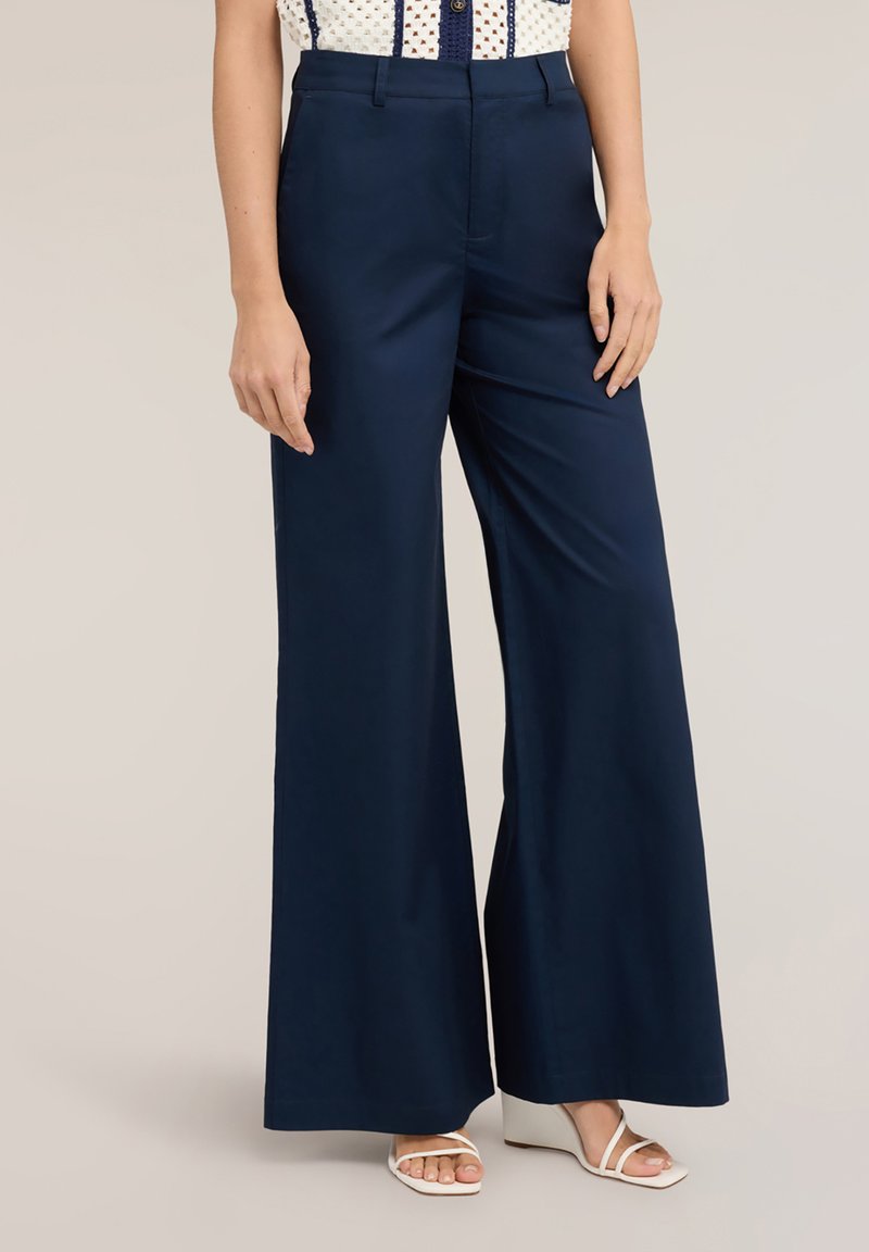 Femeie purtând pantaloni largi navy cu talie înaltă și bucle pentru curea, asortați cu sandale albe cu platformă și top cu model.