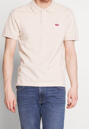Mand iført en beige poloshirt med kortærmer og en lille rød logo samt blå jeans, stående foran en ensfarvet hvid baggrund.