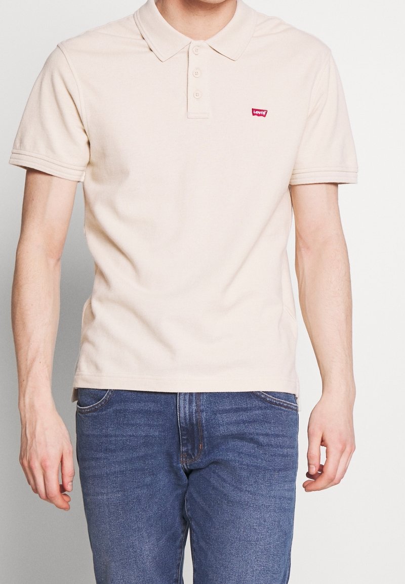 Homme portant un polo beige à manches courtes avec un petit logo rouge et un jean bleu, debout devant un fond blanc uni.