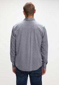 Camisa de manga larga gris con un patrón, de cuello abotonado. Presenta pequeños diseños geométricos blancos y puños con botones. La parte inferior tiene un dobladillo redondeado.