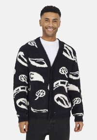 Zwart-witte fuzzy cardigan met abstracte patronen van bomen en vormen, met een sjawl kraag, lange mouwen en knopen aan de voorkant.