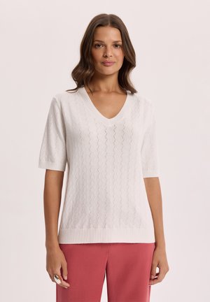 Kobieta o falowanych brązowych włosach, ubrana w biały krótki sweter z dzianiny z dekoltem w serek i różowe spodnie, stojąca na tle jednolitego tła.