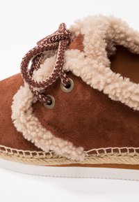 Brown suede shoe with a fluffy, cream-colored lining. Features braided burgundy laces, metal eyelets, and a woven jute midsole.