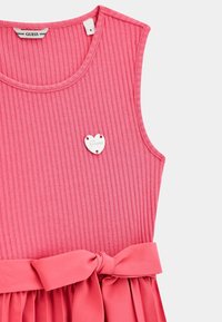 Robe sans manches rose avec une texture côtelée, un nœud à la taille et un accent en métal en forme de cœur portant le nom de la marque "Guess."