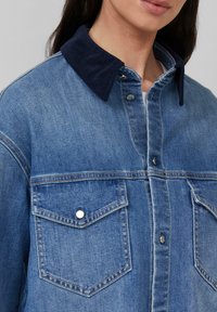 Giubbotto in denim con colletto in velluto a coste scuro, tasche sul petto anteriori e cuciture a contrasto, in un blu medio con una texture liscia.