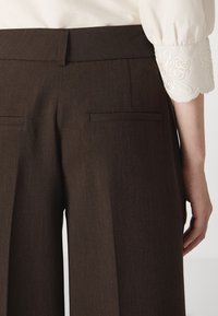 Bruine op maat gemaakte pantalons met achterzakken, gedragen met een witte lange mouw top met geborduurde golvende manchetten.