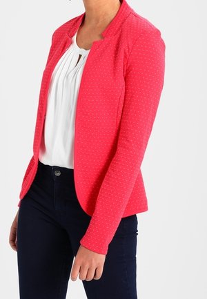 Veste rose en tissu texturé, avec de petits pois blancs, un col rond et sans fermeture visible, assortie à un pantalon bleu foncé.