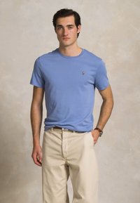 Noormees, kes kannab helesinist lühikeste varrukatega särki ja beeže pükse, seisab ühe käega taskus neutraalse tausta ees.