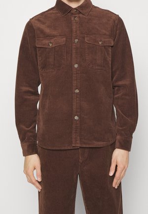 Uomo che indossa una camicia marrone in velluto a coste con due tasche sul petto e pantaloni in velluto a coste marroni abbinati.