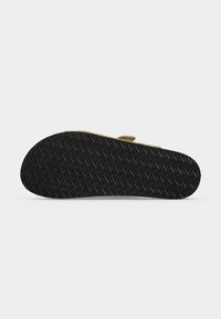Semelle de sandale avec motif de bande de roulement en zigzag noir, montrant le dessous d'une sandale beige sur un fond clair uni.