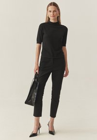 Schwarzes Strickoberteil mit kurzen Ärmeln und hohem Kragen, kombiniert mit schwarzen, maßgeschneiderten Hosen und spitzen Stilettos. Die Schultertasche ist ebenfalls schwarz.