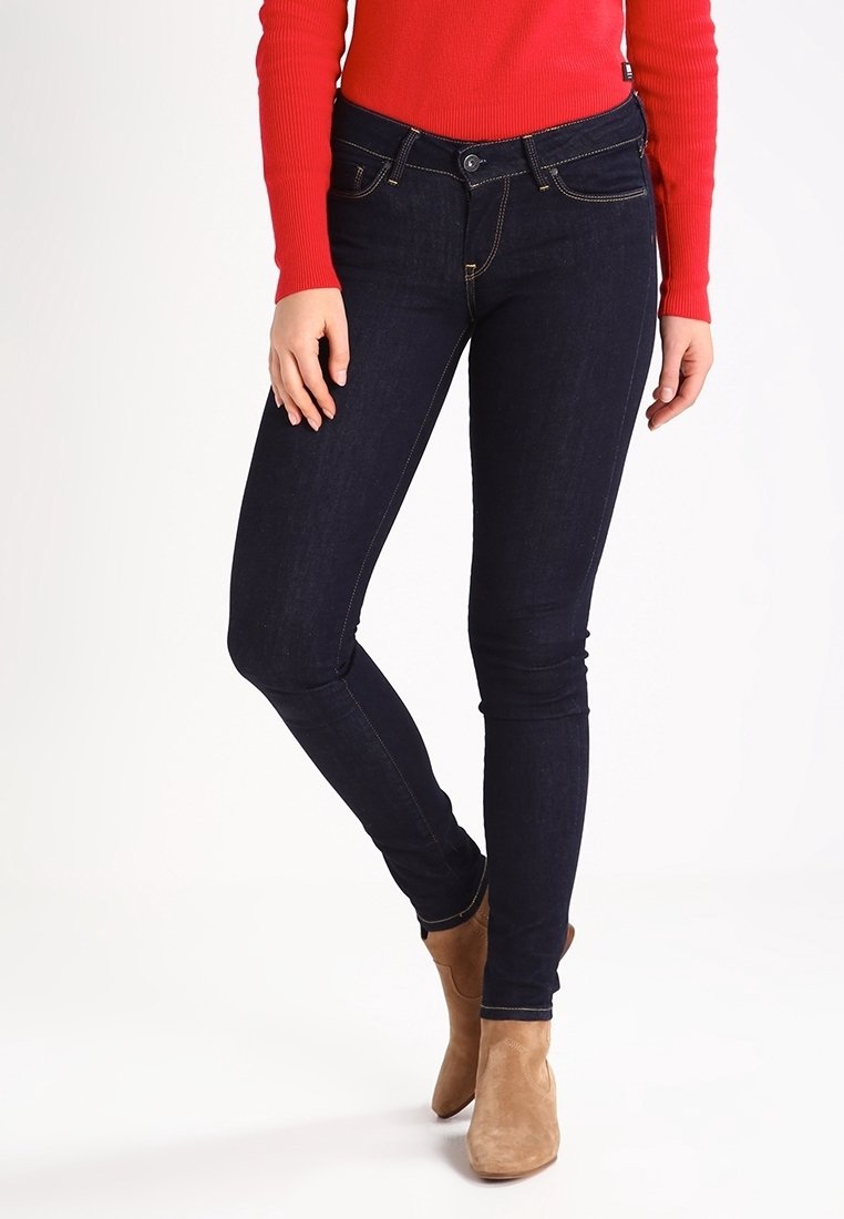 Jeans ajustados de color azul oscuro con costuras amarillas, cinco bolsillos y un cierre frontal con botón, combinados con una parte superior de manga larga de canalé roja y botas de tobillo en color marrón claro.