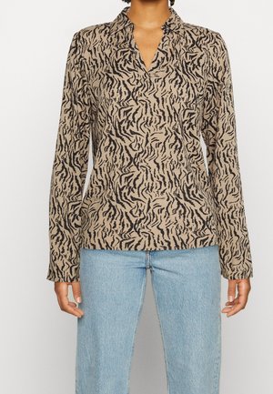 Beigen värinen pitkähiha-paita, jossa mustat abstraktit tiikerinraidat, yhdistetty vaalean sinisiin farkkuihin, kuvassa näkyy ylävartalosta lonkkiin asti.