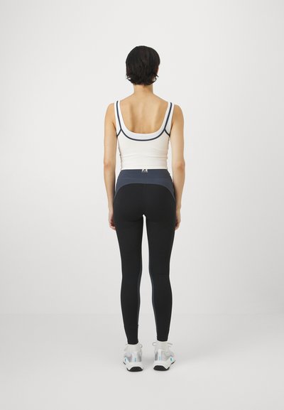 Kvinnelig idrettsantrekk med en hvit tanktop med mørke detaljer og svarte tights med blå aksent. Tightsene er tettsittende med en høy midje.