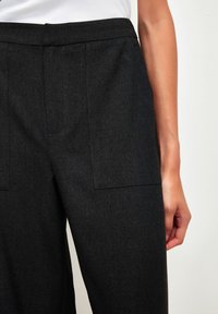 Pantalons taillés gris foncé avec une texture lisse, ceinture large et poches avant, présentés sur la moitié inférieure d'une silhouette sur un fond neutre.