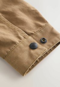 Gros plan sur le poignet d'une veste en tissu beige avec des coutures visibles et un bouton-pression noir avec du texte autour de son bord.