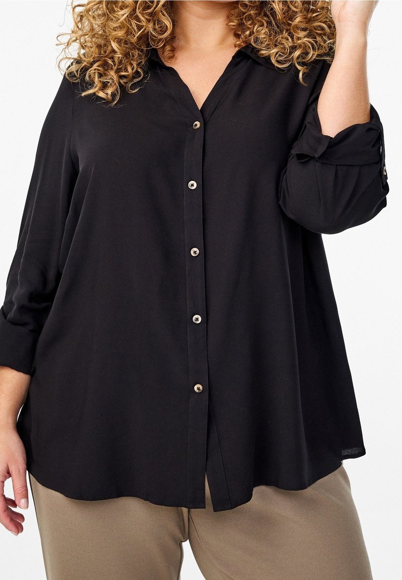 Personne aux cheveux bouclés portant une chemise noire ample boutonnée avec manches retroussées et un pantalon beige sur fond blanc.