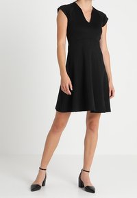 Abito nero a maniche corte con scollatura a V, corpino aderente e gonna svasata. Tessuto liscio, lunghezza al ginocchio, abbinato a tacchi a cinturino neri.