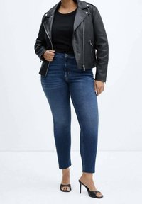 Veste de motard en cuir noir sur une chemise noire, associée à un jean bleu taille haute et des sandales à talons noirs. Textures lisses et design ajusté.